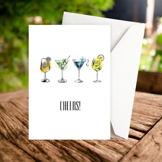 Greeting card with cocktail illustrations and 'Cheers!' text on a wooden surface with a blurred green background