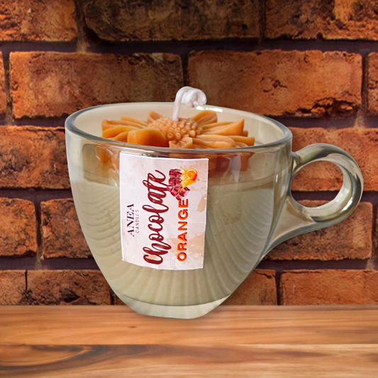 Clear mug with a floral decoration and 'ANEA' label against a brick wall. CHOCOLATE ORANGE DAISY CANDLE