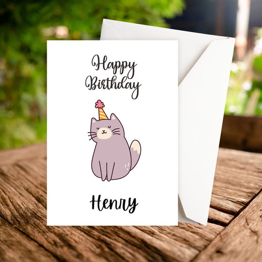 Happy Birthday personalised card with a purple cat wearing a party hat on a wooden surface with a blurred green background.