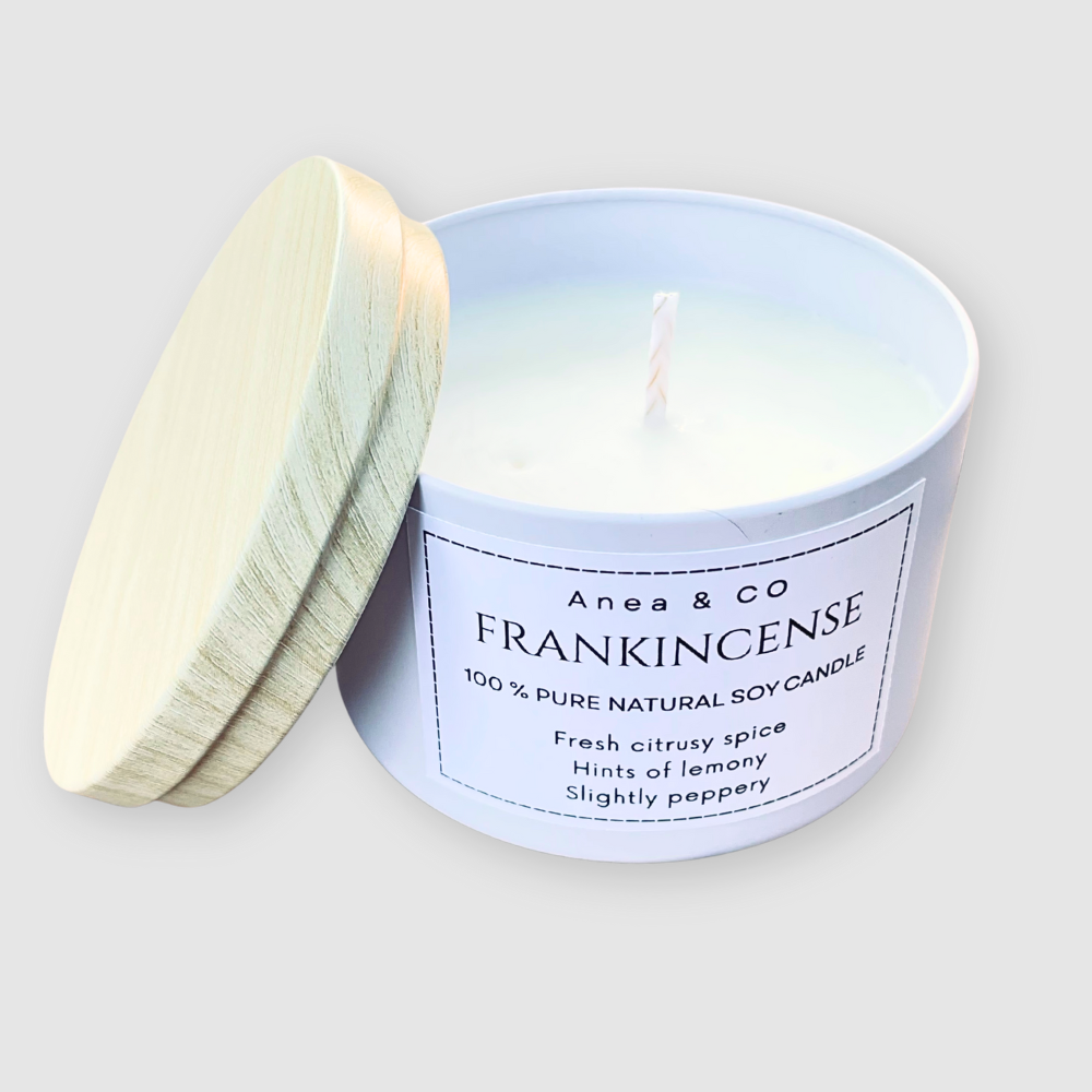 A white candle in a tin with a wooden lid, featuring a label on a light gray background. FRANKINCENSE NATURAL SOY CANDLE