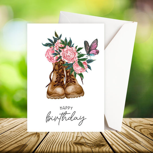 Happy Birthday card with boots, flowers, and a butterfly on a wooden surface with a blurred green background.