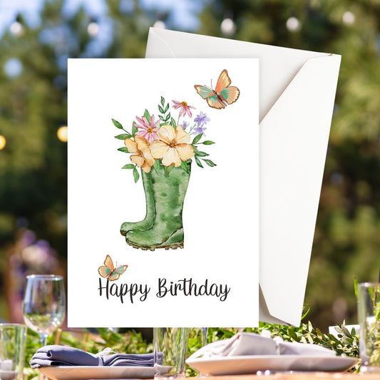 Happy Birthday card with green boot holding flowers and butterfly on a table setting outdoors
