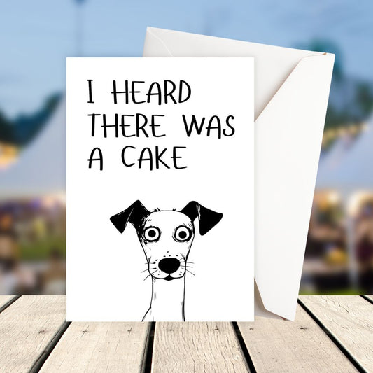 Card with a dog illustration and text 'I heard there was a cake' on a wooden surface with a blurred outdoor background. Funny Dog Cake Birthday Card