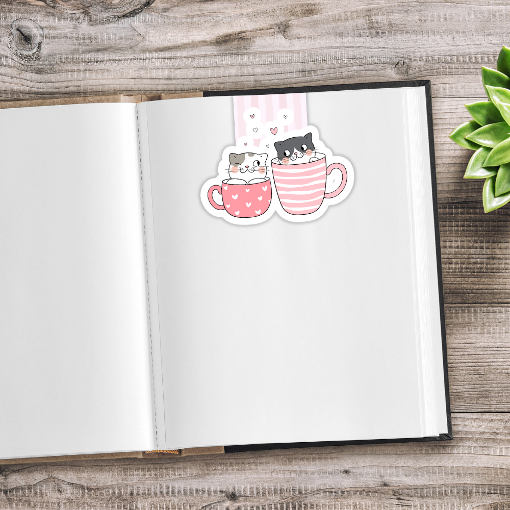 Open notebook with a sticker of two cats in a mug on a wooden surface. Kawaii Cats Magnetic Bookmark