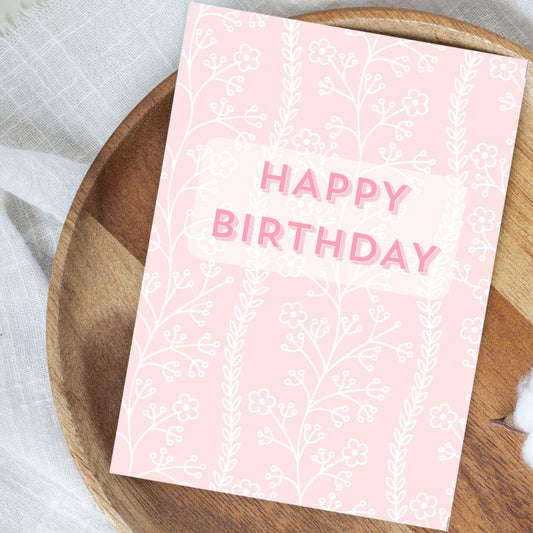 Pink birthday card with white text and floral patterns on a wooden surface. Pastel Flower Happy Birthday Card