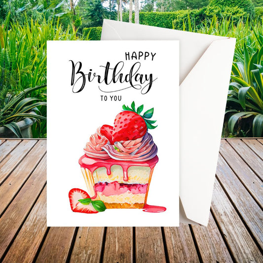 Birthday card with a strawberry cupcake illustration and 'Happy Birthday' text, placed on a wooden surface with plants in the background.