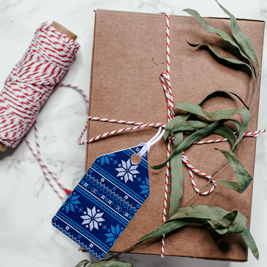 Brown gift box with red and white string, green leaves, and a blue tag on a light background. Ugly Sweater Christmas Gift Tag Set