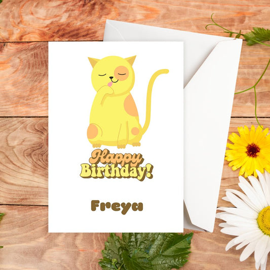 Birthday card with a yellow cat illustration and 'Happy Birthday Freya' text on a wooden surface with flowers. Yellow Cat Birthday Personalised Card