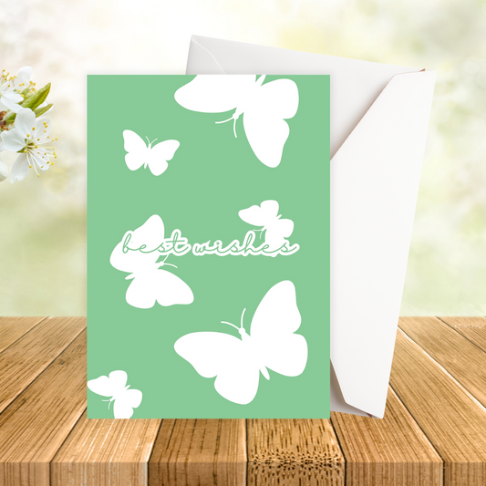 Green Butterflies Best Wishes Greeting Card with white butterflies and 'best wishes' text on a green background, placed on a wooden surface with flowers in the background.