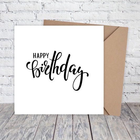 White card with 'Happy Birthday' text and brown envelope on a wooden surface with white brick wall background. HAPPY BIRTHDAY MINIMALIST CARD