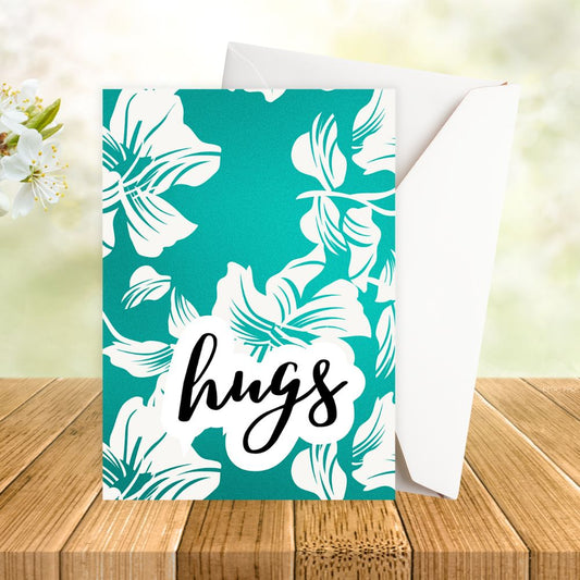 Greeting card with white floral design and 'hugs' text on a teal background, placed on a wooden surface with flowers. hugs floral happy birthday card