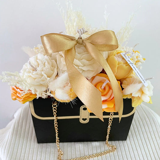 Floral arrangement in a black suitcase with a gold bow and chain on a white background. ORANGE CANDLE FLOWER BOUQUET BAG