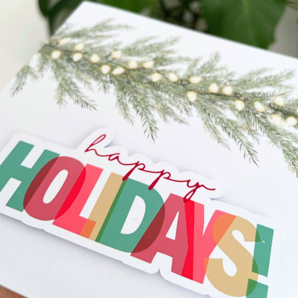 Colorful 'Happy Holidays' sign with greenery and lights on a white surface. 3D Happy Holidays Christmas Card
