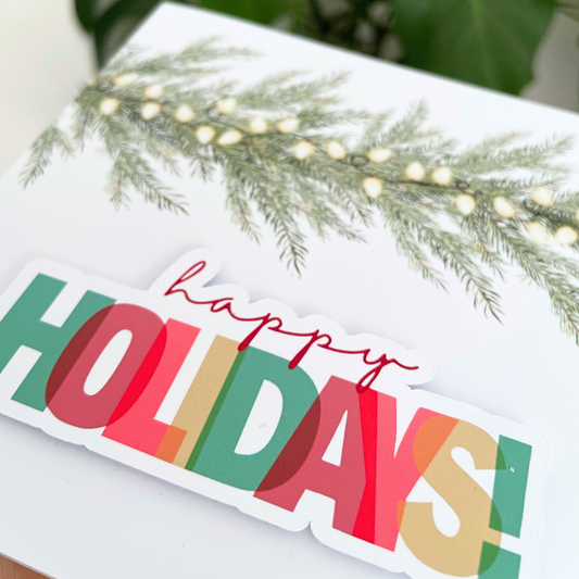 Colorful 'Happy Holidays' sign with greenery and lights on a white surface. 3D Happy Holidays Christmas Card