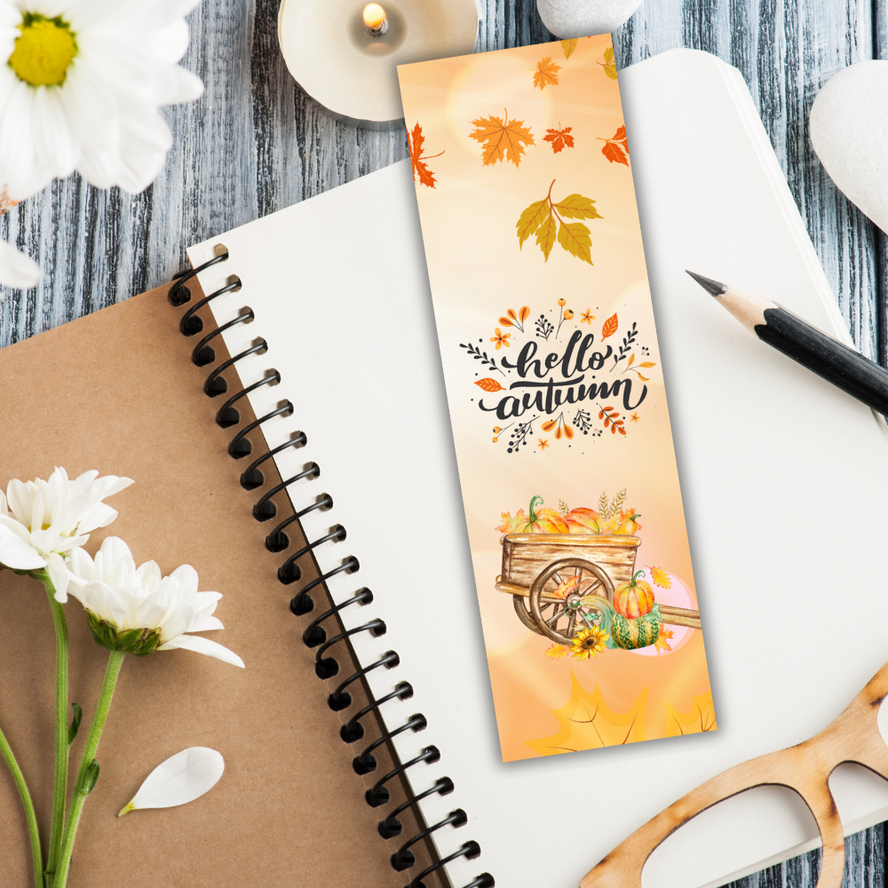 Autumn-themed bookmark with a wagon and pumpkins on a notebook with flowers and glasses.