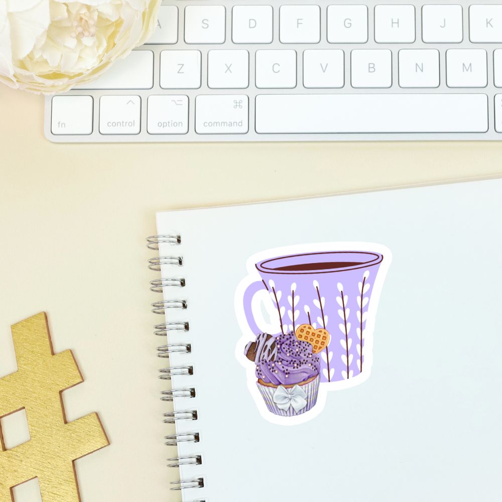 Notebook with a sticker of a purple mug and cupcake on a desk with a keyboard.Coffee and Blueberry Cupcake Sticker