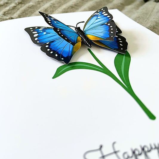 Blue butterfly on a white card with green leaves and 'Happy' text