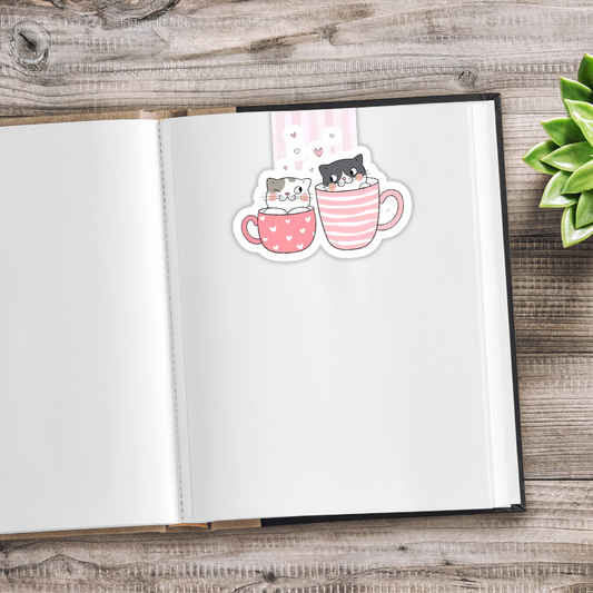 Open notebook with a sticker of two cats in a mug on a wooden surface. Kawaii Cats Magnetic Bookmark