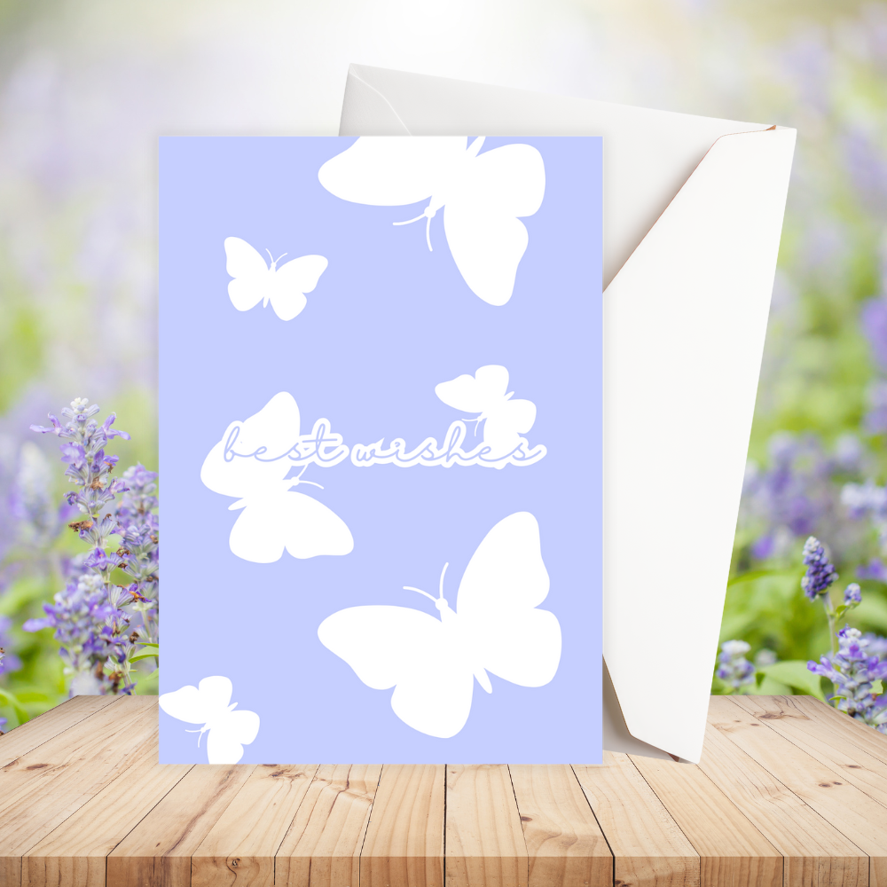 Greeting card with white butterflies and 'best wishes' text on a light blue background, placed on a wooden surface with a blurred floral background.