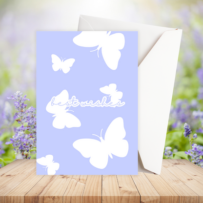 Greeting card with white butterflies and 'best wishes' text on a light blue background, placed on a wooden surface with a blurred floral background.