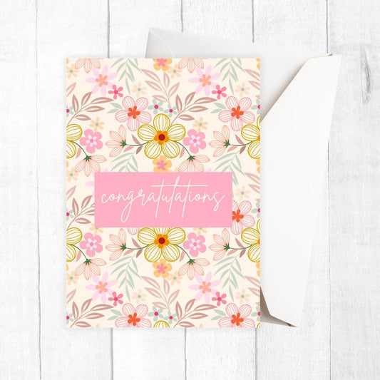 Floral congratulation card with a pink 'congratulations' text on a white wooden surface. Pink Wild Flowers Congratulations Card