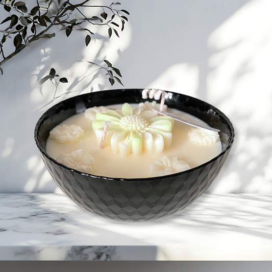 Black bowl with a flower-shaped candle on a marble surface with a plant in the background. SPRING DAISY SOY CANDLE