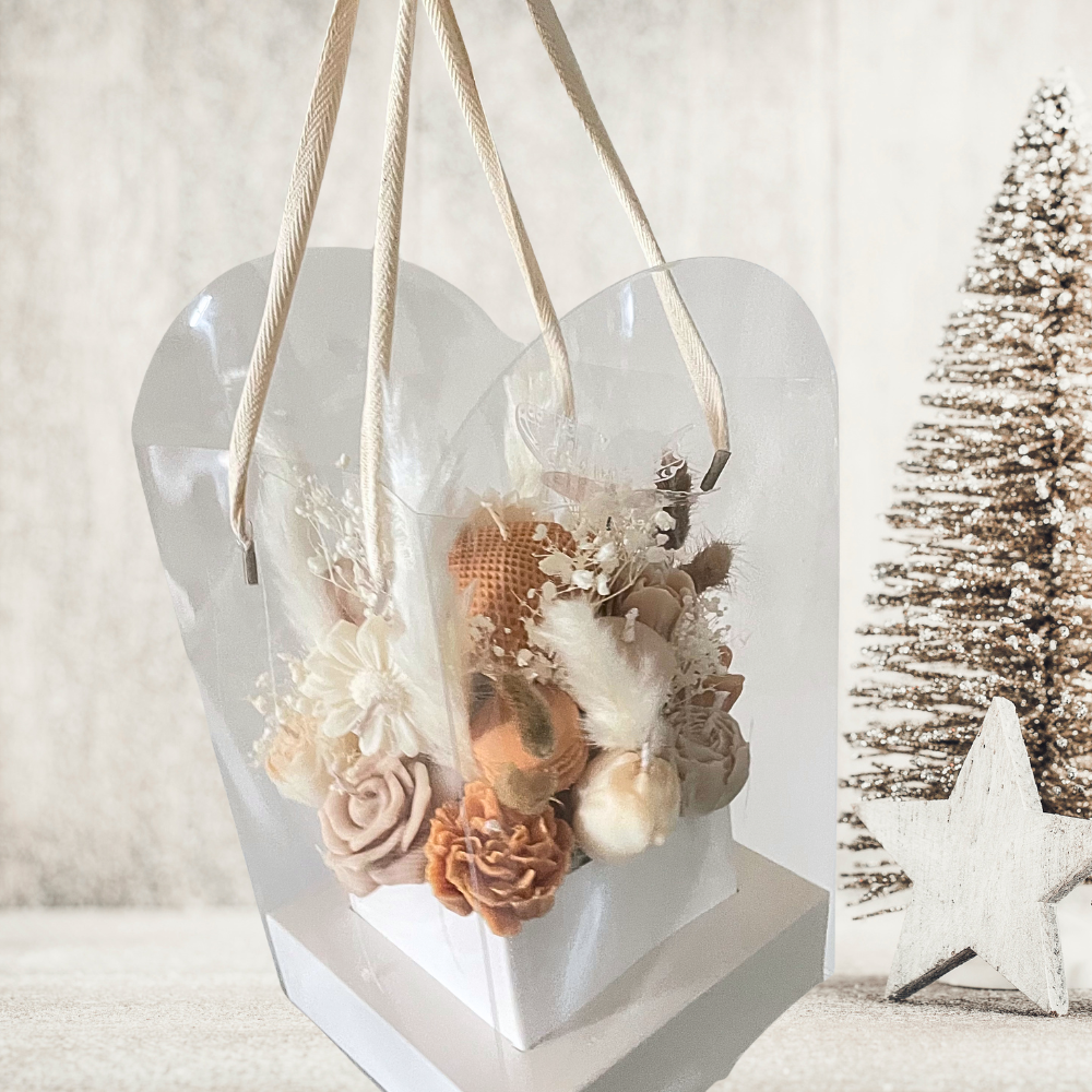 Heart-shaped transparent container with dried flowers on a white surface with a Christmas tree in the background. CHOCOLATE & CREAM CHRISTMAS CANDLE BOUQUET