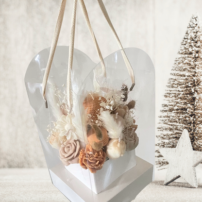 Heart-shaped transparent container with dried flowers on a white surface with a Christmas tree in the background. CHOCOLATE & CREAM CHRISTMAS CANDLE BOUQUET