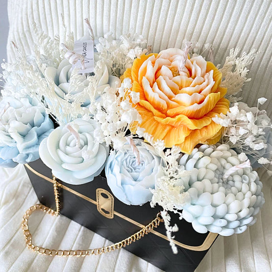 Decorative arrangement of flowers and plants in a black box with gold chain on a white textured background.BLUE CANDLE FLOWER BOUQUET BAG