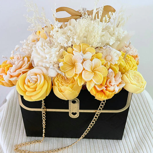 Black handbag with gold chain filled with yellow and white flowers on a white background. ORANGE CANDLE FLOWER BOUQUET BAG