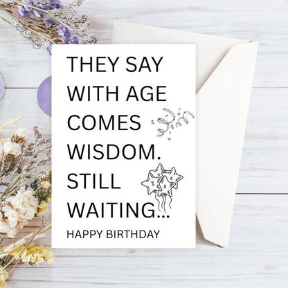 Happy Birthday card with humorous message on a wooden surface with flowers